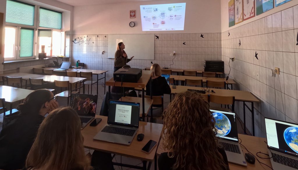 Kobieta prowadzi prezentację na temat systemów informacji geograficznej (GIS) dla grupy osób korzystających z laptopów w klasie.
