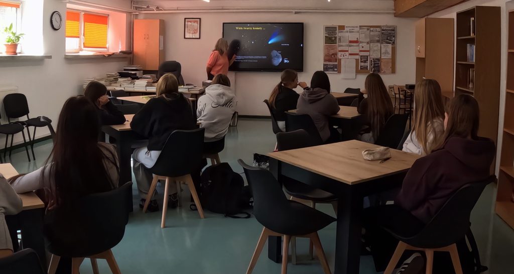 Grupa uczniów siedzi przy stolikach w sali lekcyjnej, a nauczycielka wskazuje na duży ekran z prezentacją o kometach.