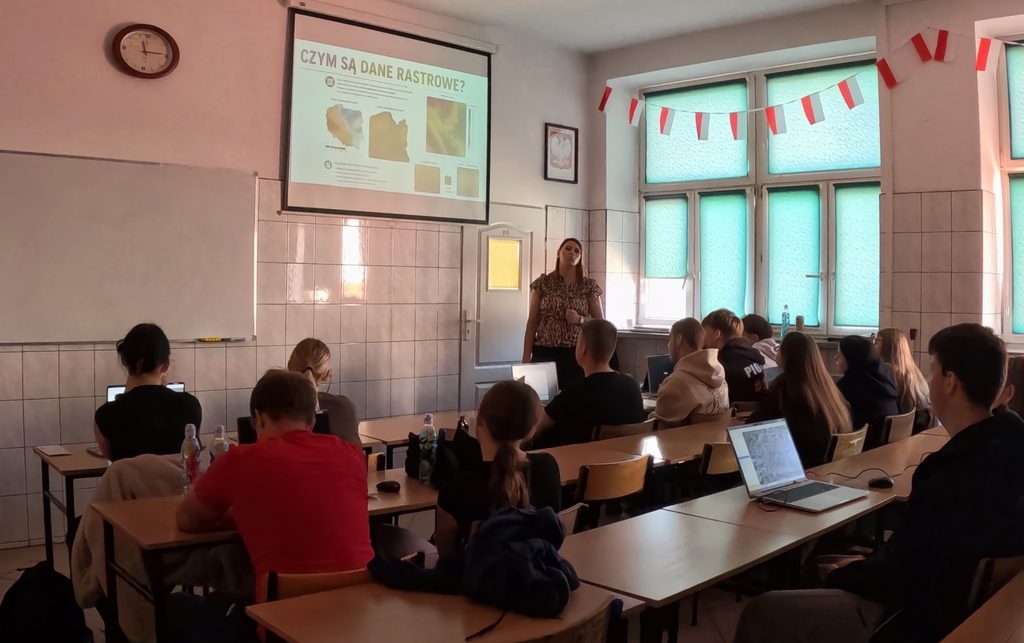 Uczniowie z laptopami słuchają nauczycielki omawiającej dane rastrowe na tablicy multimedialnej w klasie ozdobionej flagami Polski.
