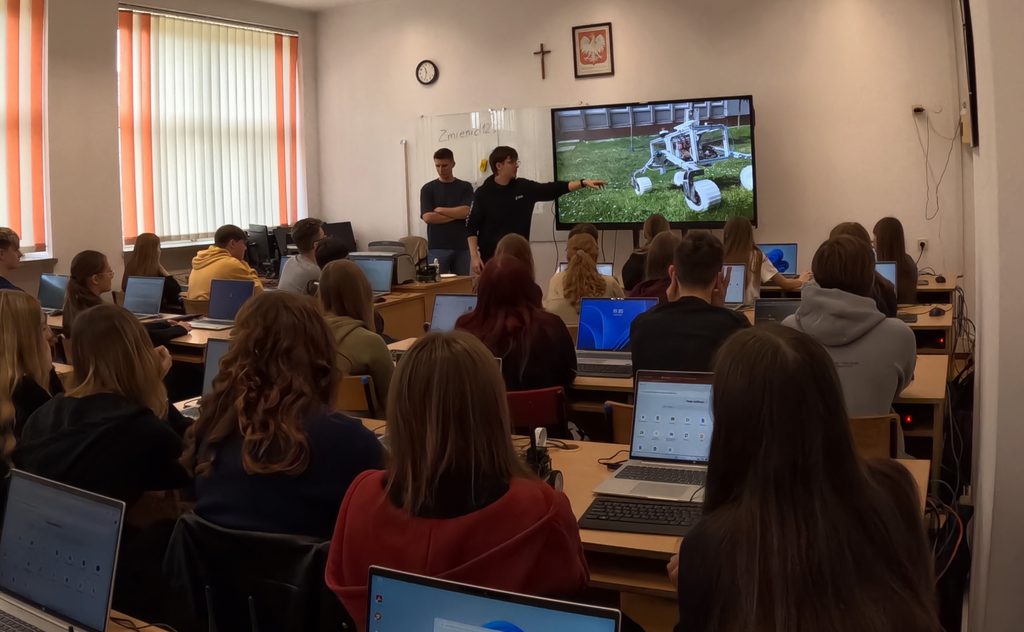 Grupa studentów podczas zajęć z komputerami, wykładowca wskazuje na ekran z modelem robota marsjańskiego.