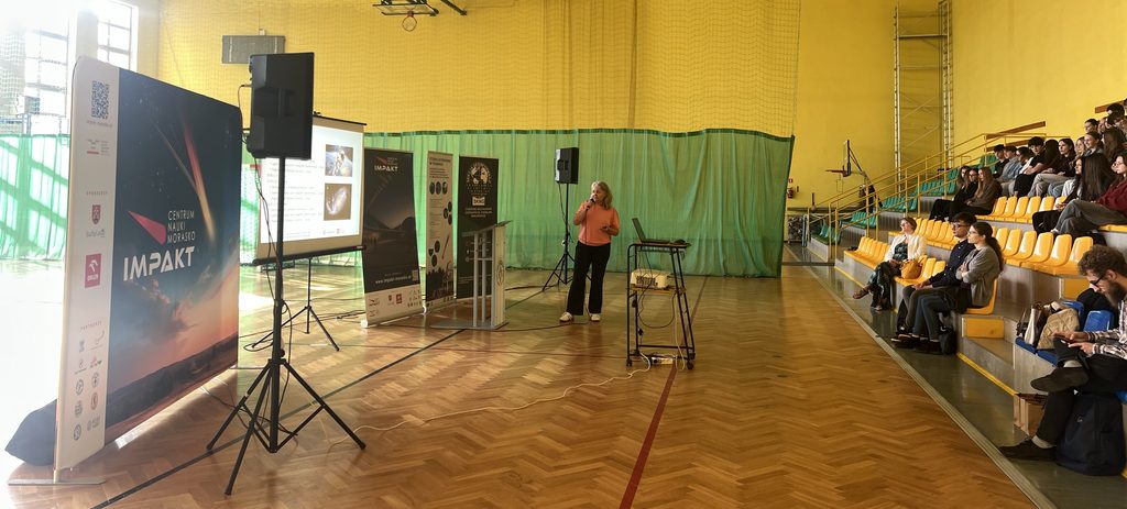 Kobieta prowadzi prelekcję przy ekranie z prezentacją, a grupa ludzi ogląda ją siedząc na trybunach w sali gimnastycznej.
