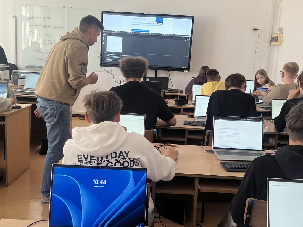 Nauczyciel pomaga uczniowi podczas lekcji informatyki, na której uczniowie pracują na laptopach, a na ekranie dużego monitora wyświetlony jest kod programistyczny.