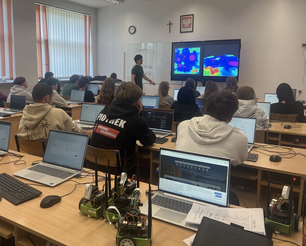 Uczniowie na lekcji informatyki pracują na laptopach, a nauczyciel pokazuje projekcję z kolorowymi mapami ciepła na dużym ekranie.