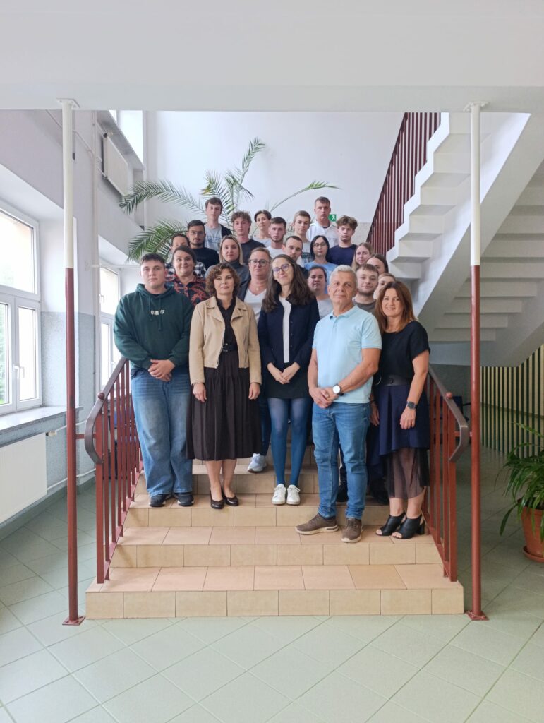 Grupa kilkudziesięciu osób w różnym wieku stoi na schodach wewnątrz budynku, w tle widoczna roślina doniczkowa.