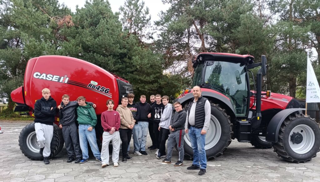 Grupa młodych mężczyzn i dorosły mężczyzna stoją obok czerwonego ciągnika rolniczego Case RB456 na tle drzew.