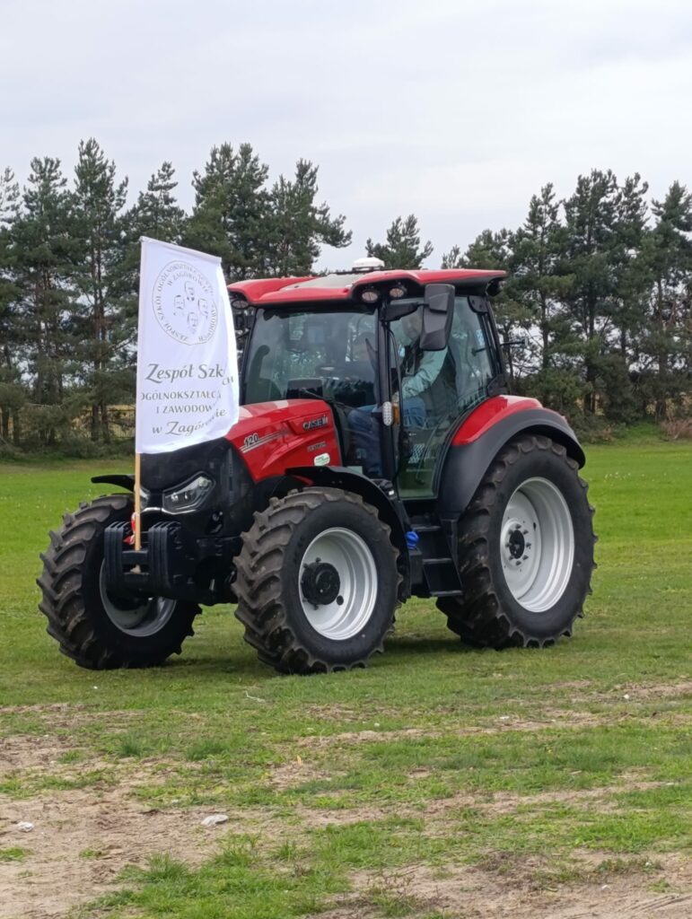 Czerwony traktor marki Case z flagą szkoły rolniczej na polu z zieloną trawą i drzewami w tle.