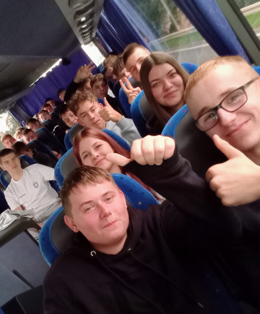 Grupa młodych osób siedzi w autobusie, niektóre pokazują kciuki w górę i uśmiechają się.