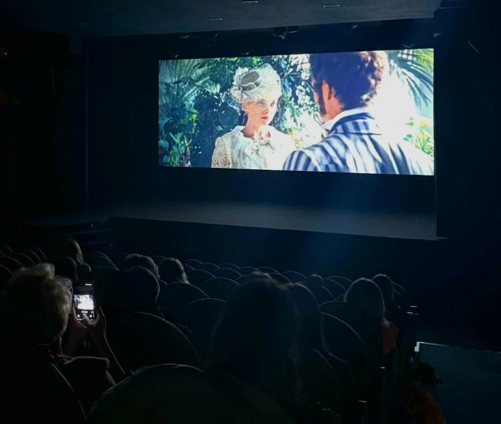 W ciemnej sali kinowej widzowie oglądają film, na ekranie kobieta w białym kapeluszu rozmawia z mężczyzną w pasiastym stroju.