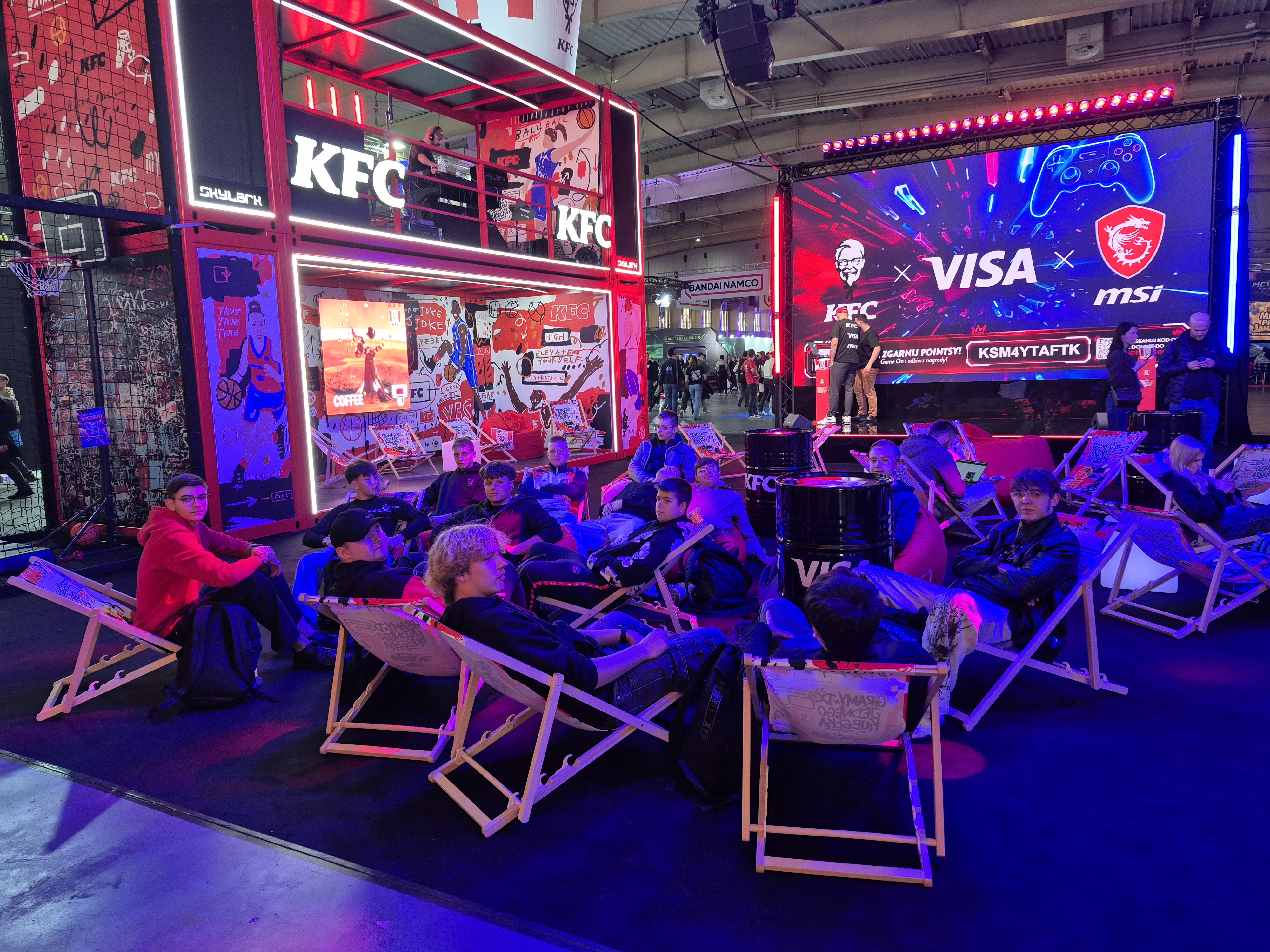 Grupa młodych osób siedzi na leżakach w strefie eventowej z dużym ekranem i neonowymi dekoracjami promującymi marki KFC, Visa i MSI.