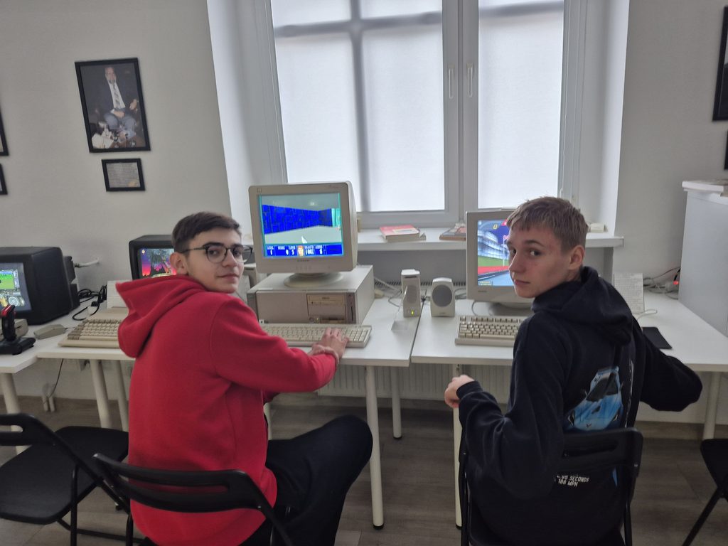Dwaj nastolatkowie grają na starych komputerach z monitorem CRT, siedząc przy białym stole w jasnym pomieszczeniu.