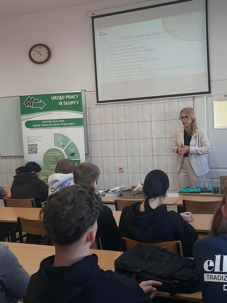 Kobieta prowadzi wykład na temat statusów osób na rynku pracy, a uczestnicy siedzą w ławkach i słuchają.