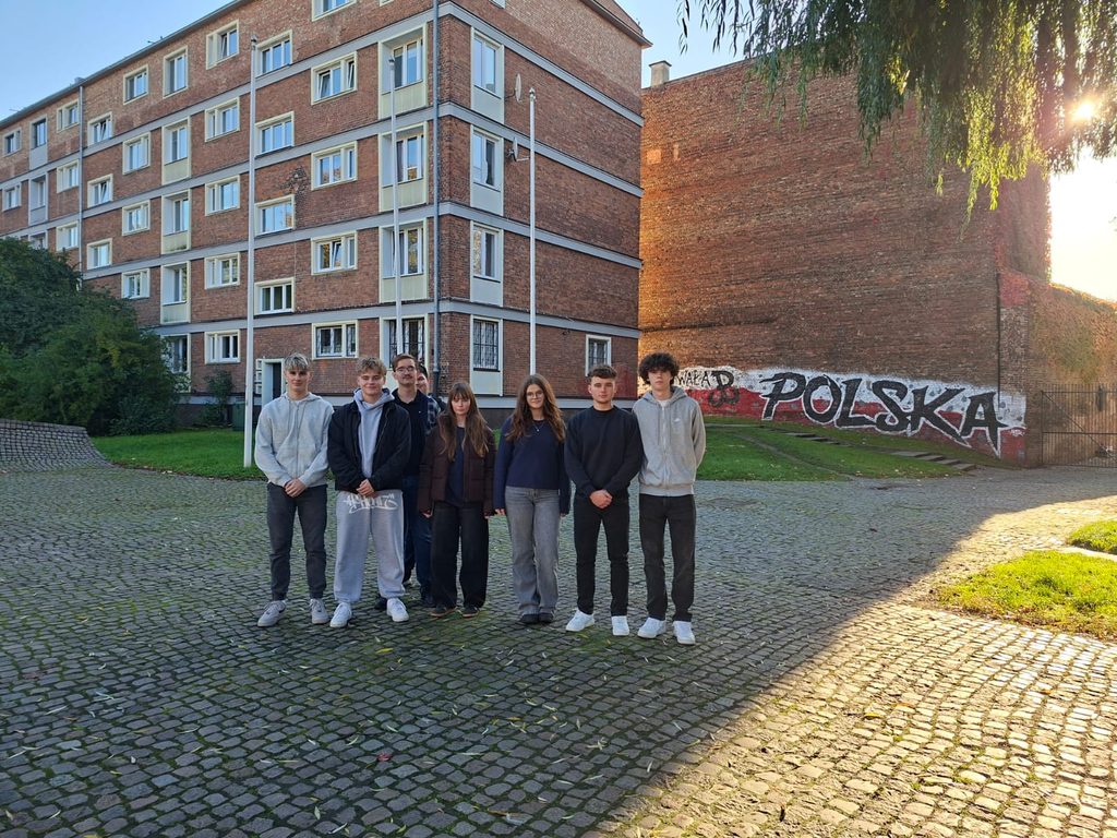 Grupa młodych ludzi stoi na brukowanym placu obok budynku mieszkalnego, na tle muru z graffiti z napisem „POLSKA”.