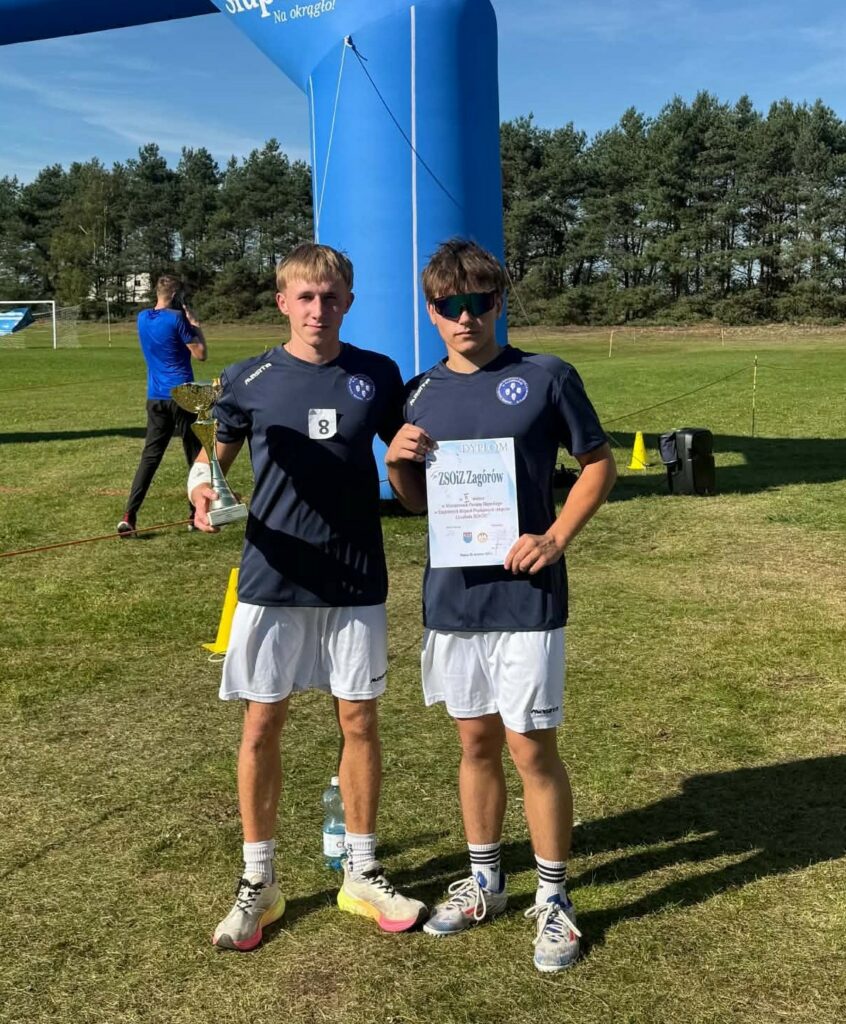 Dwaj młodzi sportowcy w strojach sportowych trzymają puchar i dyplom na tle boiska piłkarskiego i niebieskiej dmuchanej bramy.