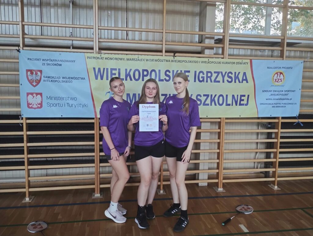 Trzy dziewczyny w fioletowych koszulkach stoją w hali sportowej, jedna z nich trzyma dyplom, a na podłodze leżą rakiety do badmintona.
