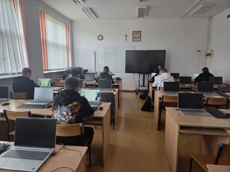 Uczniowie pracujący na laptopach w klasie informatycznej z dużym monitorem i tablicą na ścianie.
