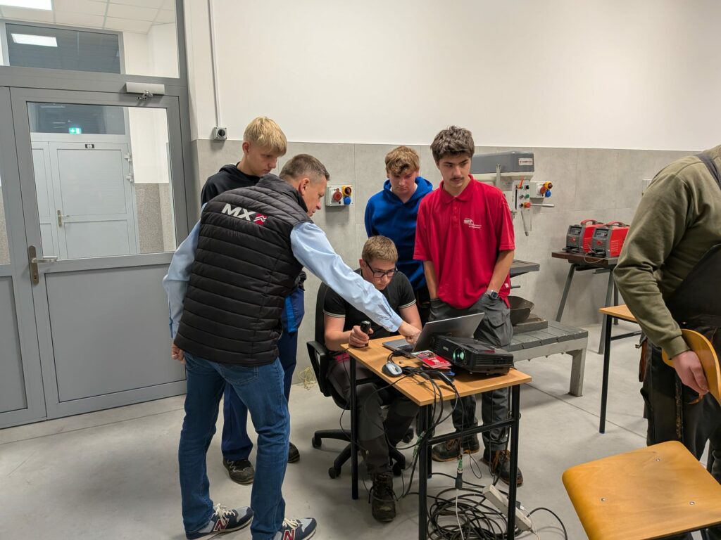 Grupa młodych mężczyzn uczy się obsługi komputera i sprzętu w warsztacie technicznym pod okiem instruktora.