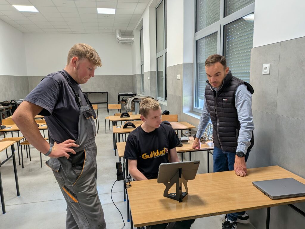 Trzech mężczyzn skupionych nad tabletem w sali lekcyjnej, jeden siedzi, dwaj stoją.