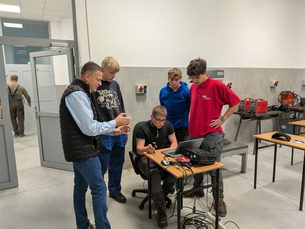 Grupa młodych mężczyzn i starszy mężczyzna obserwują pracę przy laptopie w warsztacie z narzędziami i sprzętem technicznym.
