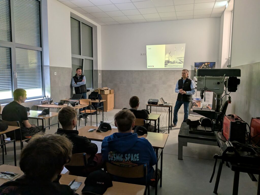 Mężczyzna prowadzi prezentację przed grupą młodych słuchaczy w sali z dużymi oknami, a na ścianie wyświetlany jest slajd z napisem „QUIZ”.